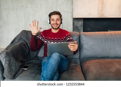 Young Smiling Man Sitting At Home In Winter, Waving Hand, Saying Hello, Wearing Red Sweater, Working On Laptop, Freelancer, Listening To Headphones, Student Studying Online, Looking In Camera