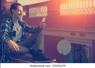 Young And Smiling Man Sitting On The Roof With Mobile Phone And Listening Music (intentional Sun Glare And Vintage Color, Focus On Mobile Phone)