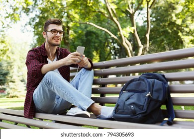 Young smiling man sitting on bench in park, listening to music with earphones on smartphone. Rest, education and relax concept, copy space