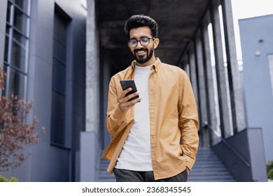 A young smiling man walks through the city with a phone in his hands, outside an office building, happily uses an application on a smartphone, reads messages, types, and browses the Internet. - Powered by Shutterstock