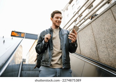 Young smiling man wearing leather jacket using mobile phone while going down escalator outdoors - Powered by Shutterstock