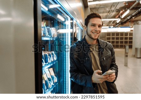 Young smiling man wearing leather jacket using mobile phone while standing near snacks vending machine in subway