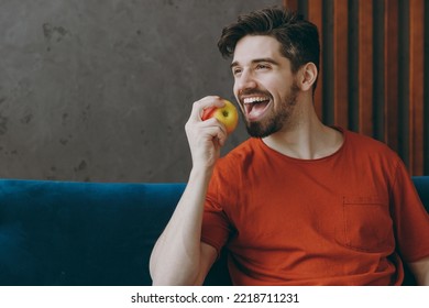 Young smiling man wears red t-shirt eat bite apple look aside sit on blue sofa couch stay at home hotel flat rest relax spend free spare time in living room indoors grey wall. People lounge concept - Powered by Shutterstock