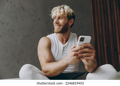 Young smiling minded fun strong sporty athletic sportsman man wear white tank shirt black shorts sit on floor hold use mobile cell phone look aside warm up training inside gym Workout sport concept - Powered by Shutterstock