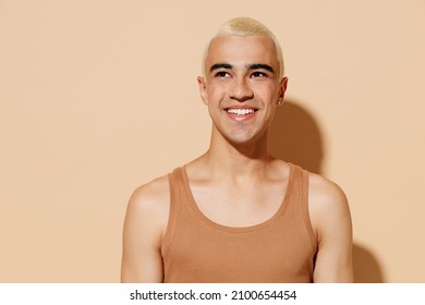 Young smiling minded laughing blond latin american gay man 20s with make up in beige tank shirt looking aside isolated on plain light ocher background studio portrait People lgbt lifestyle concept.