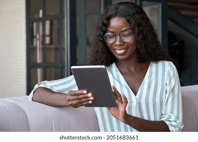 Young smiling mixed raced African black businesswoman student girl in glasses sitting on sofa holding using tablet device surfing social media apps, online chatting, ecommerce shopping at home. - Shutterstock ID 2051683661