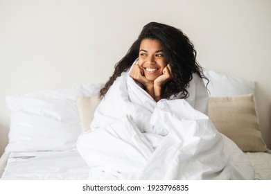 Young Smiling Mixed-race Woman With Curly Hair Placed Hands On A Chin, Covered In A Cozy White Soft Blanket Enjoying Her Day Off Or Weekend In The Warm Bed, Looking In The Window And Feeling Happy