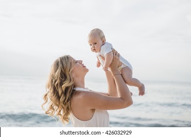 Young Smiling Mother In Flying White Dress With Her Baby On The Beach . Mother Holds Newborn Baby In Her Arms . Happy Woman With A Child Smiling And Kissing Her Baby. Close Up Portrait Out Door