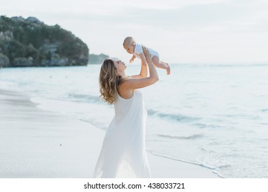 Young Smiling Mother In Flying White Dress With Her Baby On The Beach . Mother Holds Newborn Baby In Her Arms . Happy Woman With A Child Smiling And Kissing Her Baby. Close Up Portrait Out Door