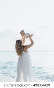 Young Smiling Mother In Flying White Dress With Her Baby On The Beach . Mother Holds Newborn Baby In Her Arms . Happy Woman With A Child Smiling And Kissing Her Baby. Close Up Portrait Out Door