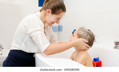 Young smiling mother pouring shampoo on hand and washing hair of her little son sitting in bath. Concept of child hygiene and health care at home. Family having time together and playing at home.
