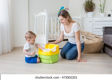 Young smiling mother sitting on floor at living room and teaching her 10 months old baby boy how to use chamber pot