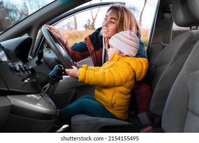 A Young Smiling Mother Teaches Her Little Child To Drive A Car. The Kid Is Sitting In A Chair And Holding The Steering Wheel With His Hands. Side View.