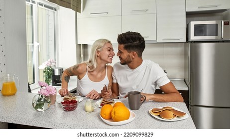 Young Smiling Multiracial Couple Looking At Each Other During Eating Pancakes On Breakfast At Table At Home. Middle Eastern Man And Blonde European Girl. Relationship And Closeness. Domestic Lifestyle