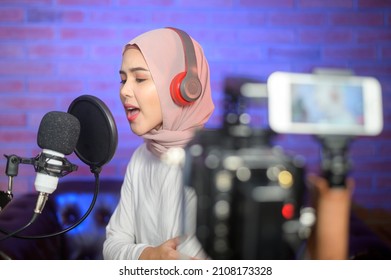 A Young Smiling Muslim Female Singer Wearing Headphones With A Microphone While Recording Song In A Music Studio With Colorful Lights.