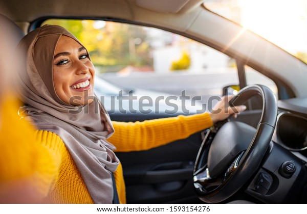 Young smiling Muslim gril making selfie portrait\
sitting in the car. Beautiful young Muslim woman in car, taking\
selfie. Road trip selfie. Young woman using her smartphone and\
making selfie