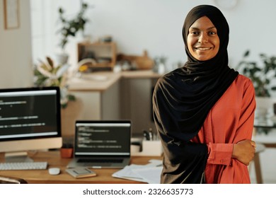 Young smiling Muslim home office worker in black hijab and red shirt looking at camera while standing against workplace with computers - Shutterstock ID 2326635773