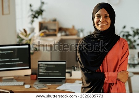 Young smiling Muslim home office worker in black hijab and red shirt looking at camera while standing against workplace with computers