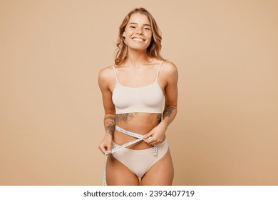 Young smiling nice lady woman with slim body perfect skin wear nude top bra lingerie stand hold measure tape on waist belly isolated on plain pastel light beige background. Lifestyle diet fit concept