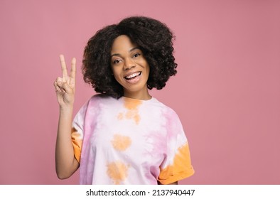 Young Smiling Nigerian Woman Showing Victory Sign Isolated On Pink Background, Copy Space. Happy Curly Haired Female  Wearing Stylish Tie Dye T Shirt Posing For Pictures In Studio
