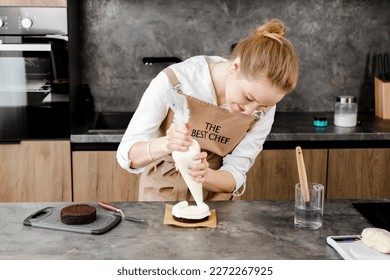 A young, smiling pastry chef in a beige apron prepares a small cake, applying frosting to a chocolate sponge cake layer through a piping bag in a modern kitchen. - Powered by Shutterstock