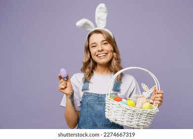 Young smiling positive fun cheerful woman wearing casual clothes bunny rabbit ears hold wicker basket show colorful egg isolated plain pastel purple background studio portrait  Happy Easter concept