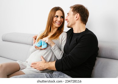 Young smiling pregnant couple relaxing on a sofa and holding baby shoes