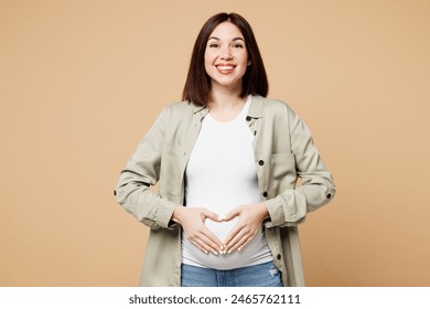 Young smiling pregnant expectant woman future mom wear grey shirt with belly stomach tummy with baby show hand heart gesture sign isolated on plain beige background. Maternity family pregnancy concept - Powered by Shutterstock