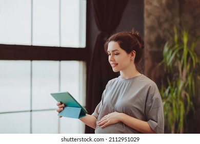 Young Smiling Pregnant Woman Holding Digital Table And Making Video Call While Sitting On Bed At Home, Talking Online With Family Or Friends. Remote Prenatal Checkup And Consultation Concept