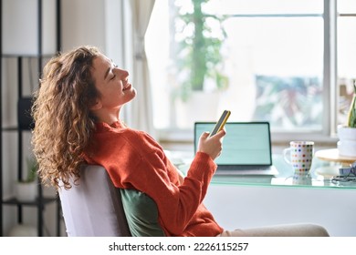 Young smiling pretty woman holding smartphone using cell mobile phone taking break relaxing while remote working or learning from home sitting on chair at table with laptop.