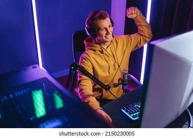 Young smiling pro cybersport gamer streaming while playing shooting game on PC at home, raising his arm, won esport tournament. Cyber sportsman enjoy his victory in competition, makes yes gesture