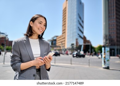 Young smiling professional Asian business woman entrepreneur wearing suit holding smartphone looking at mobile phone using apps on cellphone tech ordering taxi standing on urban city street. - Shutterstock ID 2177147489