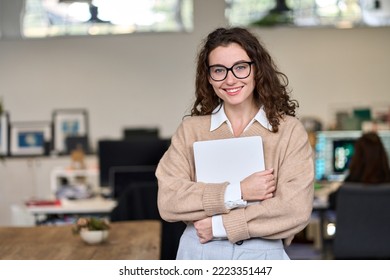 Young smiling professional business woman, happy businesswoman, female company worker intern or corporate manager holding laptop standing in modern office working, looking at camera. Portrait - Shutterstock ID 2223351447
