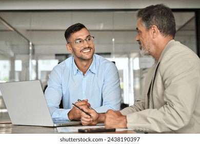 Young smiling professional latin business man working in corporate office with older executive manager using laptop technology having conversation on financial project strategy at work meeting. - Powered by Shutterstock