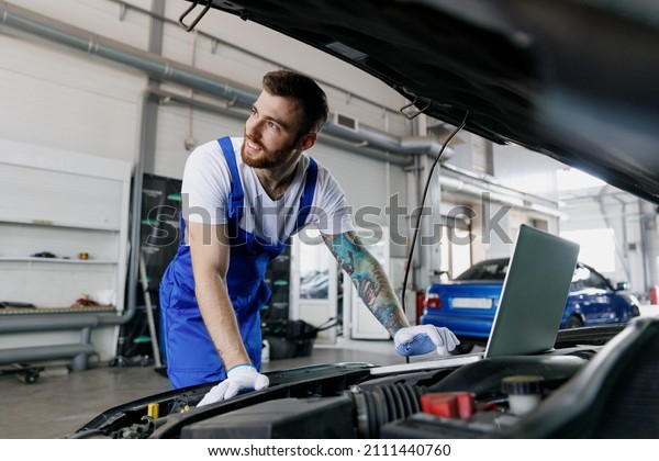 Young smiling professional technician car\
mechanic man in blue overalls t-shirt use laptop pc computer make\
diagnostics check fix problem with raised hood work in vehicle\
repair shop workshop\
indoor.