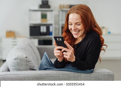 Young smiling red-haired woman using mobile phone while sitting on sofa at home - Powered by Shutterstock