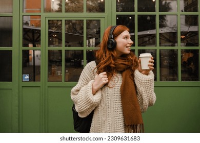 Young smiling redhead woman wearing sweater listening music with headphones and drinking coffee while standing outdoors near green door - Powered by Shutterstock