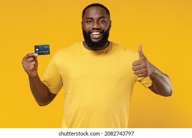 Young Smiling Rich Cool Happy Black Man 20s Wear Bright Casual T-shirt Hold In Hand Credit Bank Card Show Thumb Up Ge Isolated On Plain Yellow Color Background Studio Portrait People Lifestyle Concept