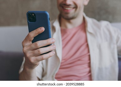 Young smiling satisfied happy man 20s wearing casual clothes beige shirt pink t-shirt use mobile cell phone in blue case sitting on grey sofa rest indoors at home on weekends. Focus on device gadget - Shutterstock ID 2039528885