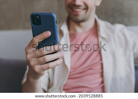 Young smiling satisfied happy man 20s wearing casual clothes beige shirt pink t-shirt use mobile cell phone in blue case sitting on grey sofa rest indoors at home on weekends. Focus on device gadget