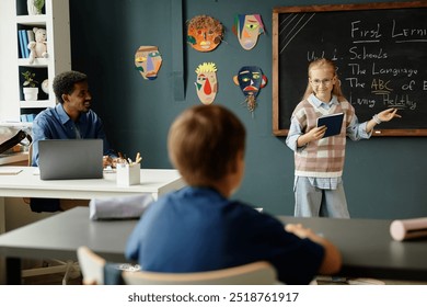 Young smiling schoolgirl pointing at blackboard during presentation answering while question from classmate in primary school classroom decorated with childrens crafts - Powered by Shutterstock