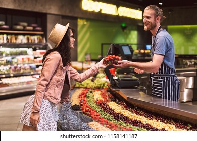 Young smiling seller in apron behind counter happily giving strawberries to customer in modern supermarket - Powered by Shutterstock