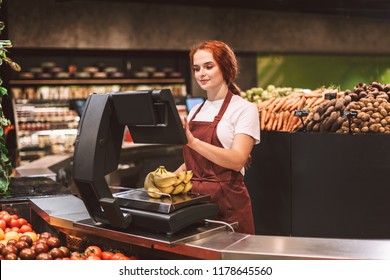Young smiling seller in apron behind counter with vegetables weighing bananas on scale in modern supermarket - Powered by Shutterstock