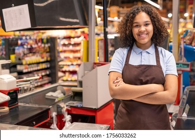 Young smiling shop assistant or cashier in workwear crossing arms by chest while standing by workplace in supermarket environment - Powered by Shutterstock