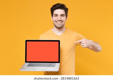 Young smiling smart IT man wear basic print design t-shirt hold use work on laptop pc computer with blank screen workspace area copy space mock up isolated on plain yellow background studio portrait
