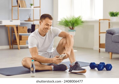 Young smiling sporty man using mobile phone after exercising sitting on yoga mat at home. Attractive guy having a rest after doing sport exercises. Healthy lifestyle and home training concept. - Powered by Shutterstock