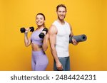 Young smiling strong fitness trainer instructor sporty two man woman wearing blue clothes spend time in home gym hold yoga mat dumbbells isolated on plain yellow background. Workout sport fit concept