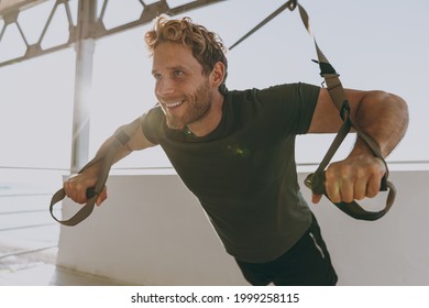 Young smiling strong sporty athletic toned fit sportsman man 20s in sports clothes warm up suspended training with TRX work out at sunrise sun dawn over sea beach seaside outdoor in summer day morning - Powered by Shutterstock