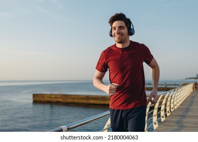 Young smiling strong sporty athletic fit sportsman man wear sports clothes heapdhones listen music warm up training at sunrise sun dawn over sea beach outdoor on pier seaside in summer day morning.