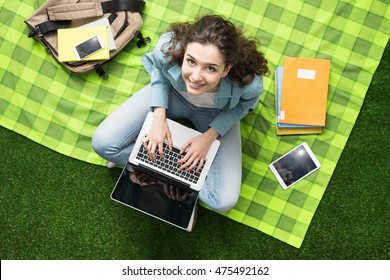 Young smiling student relaxing outdoors, she is sitting on the grass and using a laptop, summer camp concept